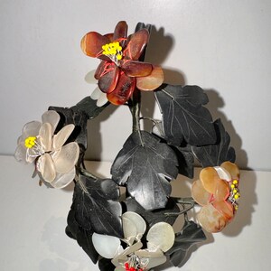 May include: A decorative floral sculpture featuring dark gray leaves and flowers in shades of brown, white, and yellow. The flowers have intricate details with yellow and red accents, creating a naturalistic aesthetic. The sculpture is likely made of stone or a similar material.