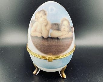 Puede incluir: Una caja de porcelana en forma de huevo con un diseño azul y blanco. La caja presenta dos querubines con alas, pintados en un estilo vintage. La caja tiene un borde dorado y tres patas doradas.