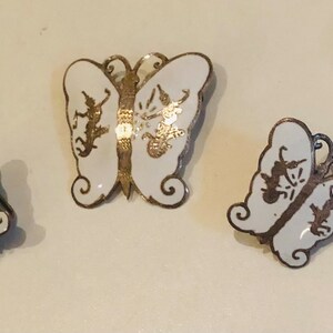 May include: A set of three silver butterfly earrings with white enamel and gold accents. The butterflies have a delicate design with intricate details.