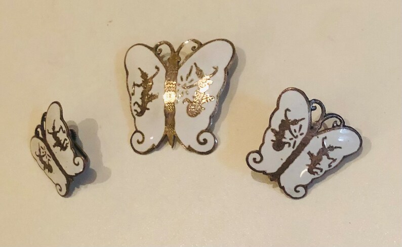 May include: A set of three silver butterfly earrings with white enamel and gold accents. The butterflies have a delicate design with intricate details.
