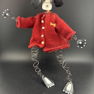 May include: A whimsical dog ornament with a black and white head, black ears, and a red sweater. The dog has wire arms and legs, with polka-dot hands and feet. A gold-colored bone-shaped accent is on the sweater. The ornament hangs from a gold string.