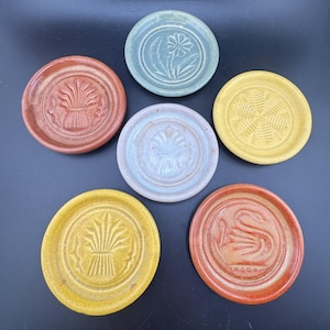 May include: A collection of six ceramic coasters in various colors, including red, yellow, and blue. Each coaster features a unique embossed design, such as a flower, wheat, or a swan. The coasters are round and appear to be handmade.