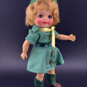 Könnte beinhalten: Vintage-Puppe mit blondem Haar, blauen Augen und rosigen Wangen. Die Puppe trägt ein grünes Kleid mit gelber Krawatte, einen passenden grünen Hut und braune Schuhe. Die Puppe ist etwa 25 cm groß.