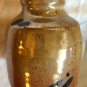 Puede incluir: Una jarra de cerámica marrón con una boca ancha y un esmalte brillante. La jarra tiene manchas negras y un diseño negro en la parte inferior.