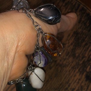 Pode incluir: Um bracelete de corrente de prata com sete pedras de diferentes cores envoltas em arame. As pedras s&atilde;o cinzas, pretas, &acirc;mbar, roxas, brancas e verdes.