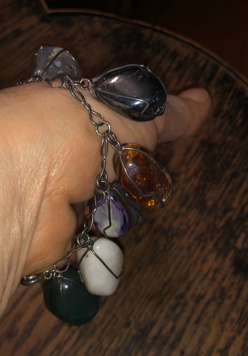 Pode incluir: Um bracelete de corrente de prata com sete pedras de diferentes cores envoltas em arame. As pedras s&atilde;o cinzas, pretas, &acirc;mbar, roxas, brancas e verdes.