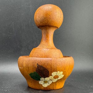 Könnte beinhalten: Ein Mörser mit Stößel aus Holz mit hellbrauner Maserung. Die Außenseite der Schale ist mit einem gemalten Motiv aus zwei weißen Blüten mit gelben Mitten und einem dunkelbraunen Blatt verziert, das die natürliche Holzstruktur hervorhebt.