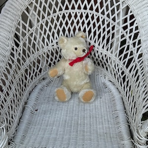 Puede incluir: Una silla de mimbre blanca con un oso de peluche blanco con una cinta roja sentado en el asiento.