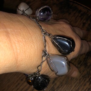 Pode incluir: Um bracelete de corrente de prata com seis pedras de diferentes cores envoltas em arame. As pedras s&atilde;o ametista, quartzo transparente, &ocirc;nix preto, &aacute;gata cinza e outras duas pedras.