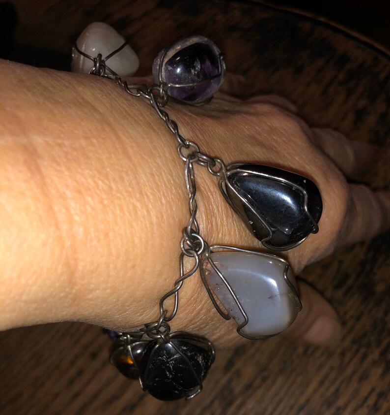 Pode incluir: Um bracelete de corrente de prata com seis pedras de diferentes cores envoltas em arame. As pedras s&atilde;o ametista, quartzo transparente, &ocirc;nix preto, &aacute;gata cinza e outras duas pedras.