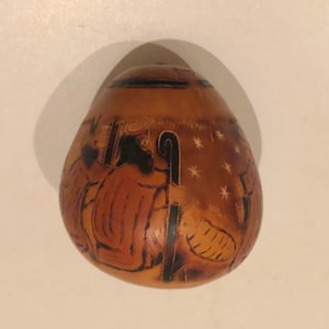 Può includere: Una zucca marrone con una scena della natività scolpita. La scena rappresenta un pastore con un bastone, una pecora e una stella.