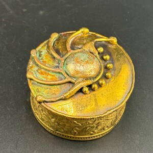 Può includere: Una piccola scatola rotonda in ottone antico con un design floreale in rilievo dettagliato sulla base. Il coperchio presenta un design ornato in rilievo con un ovale centrale e accenti decorativi. La scatola ha un aspetto invecchiato.