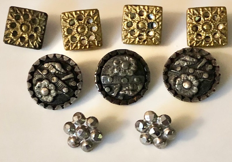 May include: Assortment of vintage buttons in gold and silver tones. The buttons feature various designs, including floral motifs, geometric patterns, and a raised design.