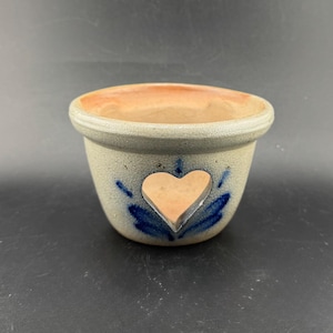 Puede incluir: Pequeño cuenco redondo de gres, con exterior gris claro e interior marrón claro natural. Un recorte en forma de corazón está centrado en el frente, con detalles pintados en azul. El cuenco probablemente se utiliza con fines decorativos.