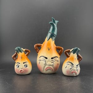 May include: A vintage ceramic salt and pepper shaker set featuring anthropomorphic onion-shaped figures with expressive faces. The set includes a tall vinegar bottle and two smaller shakers, one marked "S" and the other "P". The vinegar bottle is marked with a "V".