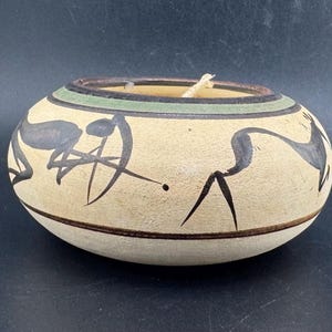 Op de afbeelding: Een ronde, beige keramische kandelaar met zwarte dierenfiguren rond de buitenkant geschilderd. De kandelaar heeft een groene en bruine band rond de bovenrand. Er staat een witte kaars in.