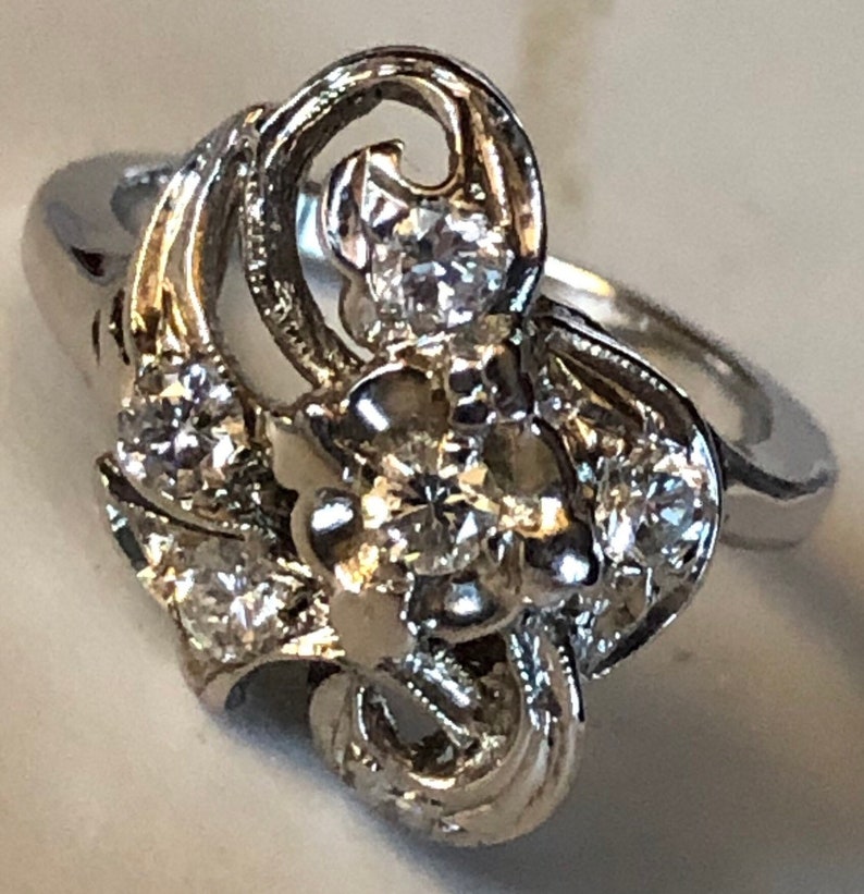 May include: A silver ring with a delicate, intricate design featuring multiple small diamonds. The ring has a central diamond surrounded by smaller diamonds in a floral pattern.