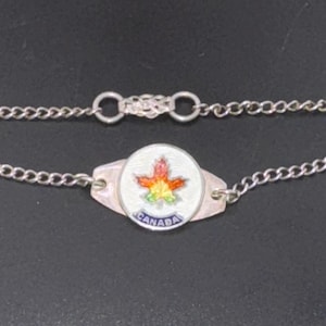 May include: Silver-toned bracelet featuring a central charm with a colorful maple leaf design and the word "CANADA" in blue. The bracelet has a chain link design and a clasp.