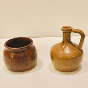 May include: Two brown ceramic pottery pieces. One is a small, round jar with a wide opening. The other is a small jug with a handle and a rounded body. Both pieces have a rustic, handmade appearance.