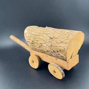 May include: A wooden toy wagon with a log for a bed. The wagon has two wheels and a wooden handle. The log is made of natural wood with bark still attached.