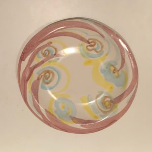 Puede incluir: Un tazón de cerámica blanca con un diseño de espiral rosa, amarillo y azul. El tazón está decorado con un patrón repetitivo de espirales y puntos.
