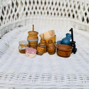 Puede incluir: Una colección de artículos de cocina en miniatura de madera, que incluyen una batidora de mantequilla, un tarro de jalea, un tarro de mantequilla de manzana, un cubo de madera, un tazón de madera, una jarra de madera y una estufa pequeña. Los artículos están pintados en varios tonos de marrón, azul y rosa.