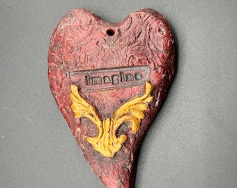 Könnte beinhalten: Ein rotes herzförmiges Ornament mit einem goldenen Design und dem Wort "Imagine" darauf.