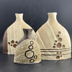 May include: Three ceramic vases in a neutral color palette. The vases have a textured, organic design with brown accents and circular patterns. The tallest vases have a bottle-like shape, while the smallest is more rounded.