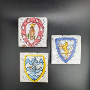 Può includere: Tre piastrelle quadrate in ceramica con finitura smaltata bianca craquelé. Ogni piastrella presenta un disegno di scudo araldico dipinto a mano: un gufo marrone con corona, un unicorno giallo e una creatura marina blu su linee ondulate.