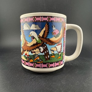 Puede incluir: Taza de cerámica blanca con un colorido diseño nativo americano. La taza presenta un borde geométrico rosa y marrón, y una ilustración detallada de figuras con alas de águila. El fondo es azul con nubes y hierba verde.
