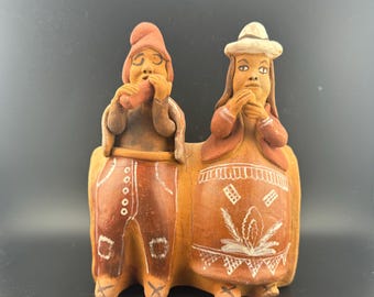 May include: A pair of hand-painted terracotta figurines depicting a man and a woman in traditional clothing. The man is playing a flute and the woman is holding her hands together. The figurines are brown with white details.
