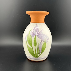 Può includere: Un piccolo vaso in ceramica bianca con un bordo e una base in terracotta. Il vaso è decorato con un fiore di iris viola dipinto a mano con foglie verdi.