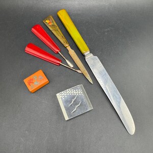 May include: A collection of vintage items including a long knife with a yellow handle, a metal nail file with a fruit-patterned handle, two red-handled metal tools, an orange rectangular tile with floral art, and a clear square block with bird icons.