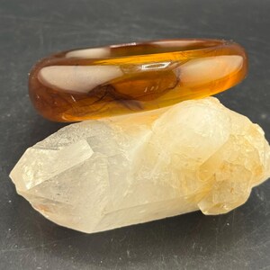 Puede incluir: Una pulsera de brazalete translúcida de color ámbar descansa sobre un cristal de cuarzo rugoso de color amarillo claro. La pulsera tiene una forma lisa y redondeada. El cristal de cuarzo tiene una forma natural e irregular, con facetas visibles y un aspecto ligeramente turbio.