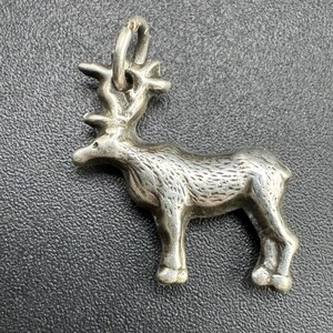 May include: A silver-toned metal charm shaped like a standing moose or reindeer, featuring detailed textured fur markings on the body and a prominent set of antlers. A small circular jump ring is attached to the top of the antlers for hanging.