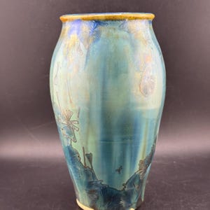 May include: A ceramic vase with a crystalline glaze featuring shades of teal, blue, and green. The surface displays intricate, flower-like crystal formations in darker blue and gold tones, with a smooth, rounded body and a distinct yellow-brown rim.