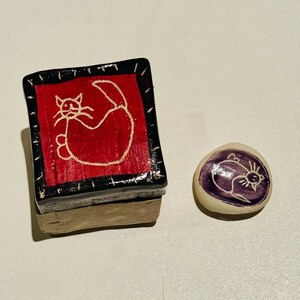 May include: Two small ceramic cat figurines. One is a red box with a black border and a white outline of a cat on the lid. The other is a round, white figurine with a purple cat on it.