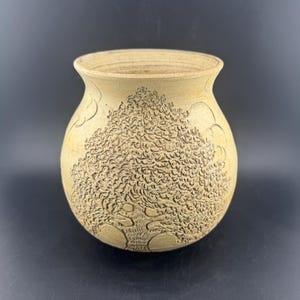 May include: A beige ceramic vase with a detailed tree design carved into the surface. The tree's branches and leaves are intricately rendered, and the vase has a rounded shape with a slightly flared rim. The background is a solid black.