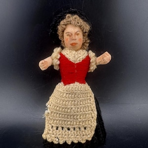 Op de afbeelding: Een vintage pop met een geschilderd gezicht, krullend haar en een zwarte hoed. De pop draagt een rood vest, een crèmekleurige gehaakte rok en een zwarte rok. De armen van de pop zijn uitgestrekt.
