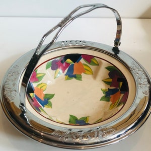 Puede incluir: Un bol de servir plateado con asa. El bol tiene un fondo blanco con un patrón floral colorido.