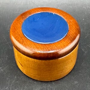 May include: A small, round wooden box with a dark brown rim and a bright blue center. The box has a natural wood grain finish on the sides and a glossy top. The design is simple and elegant, suitable for storing small items.