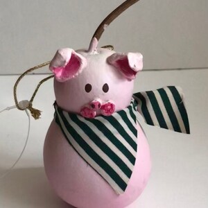 May include: A pink pig ornament with black eyes and a green and white striped scarf. The ornament has a gold string for hanging.
