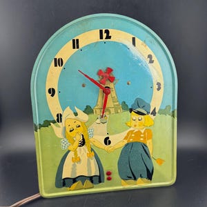 May include: Vintage children's clock with a light blue face and a whimsical design. The clock features a scene with two cartoon figures, a windmill, and a red clock hand. The clock is made of metal and has a brown power cord.