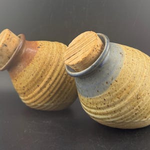 Puede incluir: Dos tarros de cerámica hechos a mano con tapones de corcho. Los tarros son de color beige con una textura acanalada y un borde marrón y azul. Los tapones de corcho son de color marrón claro. Los tarros están sobre una superficie oscura.