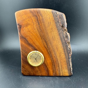 May include: A handcrafted wooden clock with a natural edge. The clock features a small, round, gold-colored clock face with black numerals and hands. The wood has a rich, warm brown color.