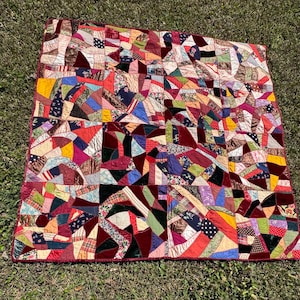 Puede incluir: Un edredón de retazos colorido con una variedad de patrones y texturas de tela. El edredón presenta una mezcla de formas geométricas en tonos de rojo, azul, amarillo y verde. El edredón está colocado sobre una superficie cubierta de hierba.