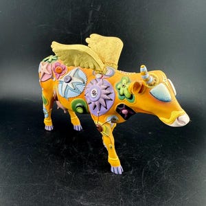 May include: A whimsical, orange-yellow cow figurine with decorative floral and geometric patterns. The cow has gold-colored wings and multi-colored accents, including blue, pink, and green. The cow's horns are striped, and its hooves are purple.
