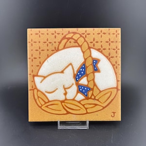 Puede incluir: Un azulejo de cerámica con un gato blanco durmiendo en una cesta. El gato tiene una cinta azul con lunares blancos alrededor del cuello. La cesta está hecha de cuerda marrón y tiene un fondo marrón y naranja. El azulejo tiene un fondo marrón y naranja con un patrón de pequeñas cruces blancas. El azulejo está firmado con la letra "J".