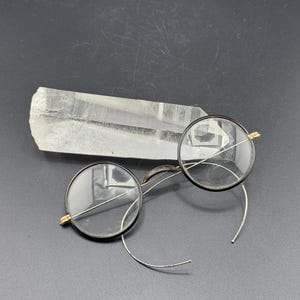 Puede incluir: Un par de gafas redondas con montura negra y patillas de alambre plateado. Las gafas están sobre un cristal de cuarzo transparente.