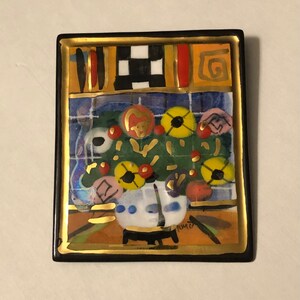 May include: A square ceramic tile with a black frame and gold trim. The tile features a colorful painting of a vase with flowers in shades of yellow, red, and blue. The artist's signature, "Tomz", is visible in the lower right corner.